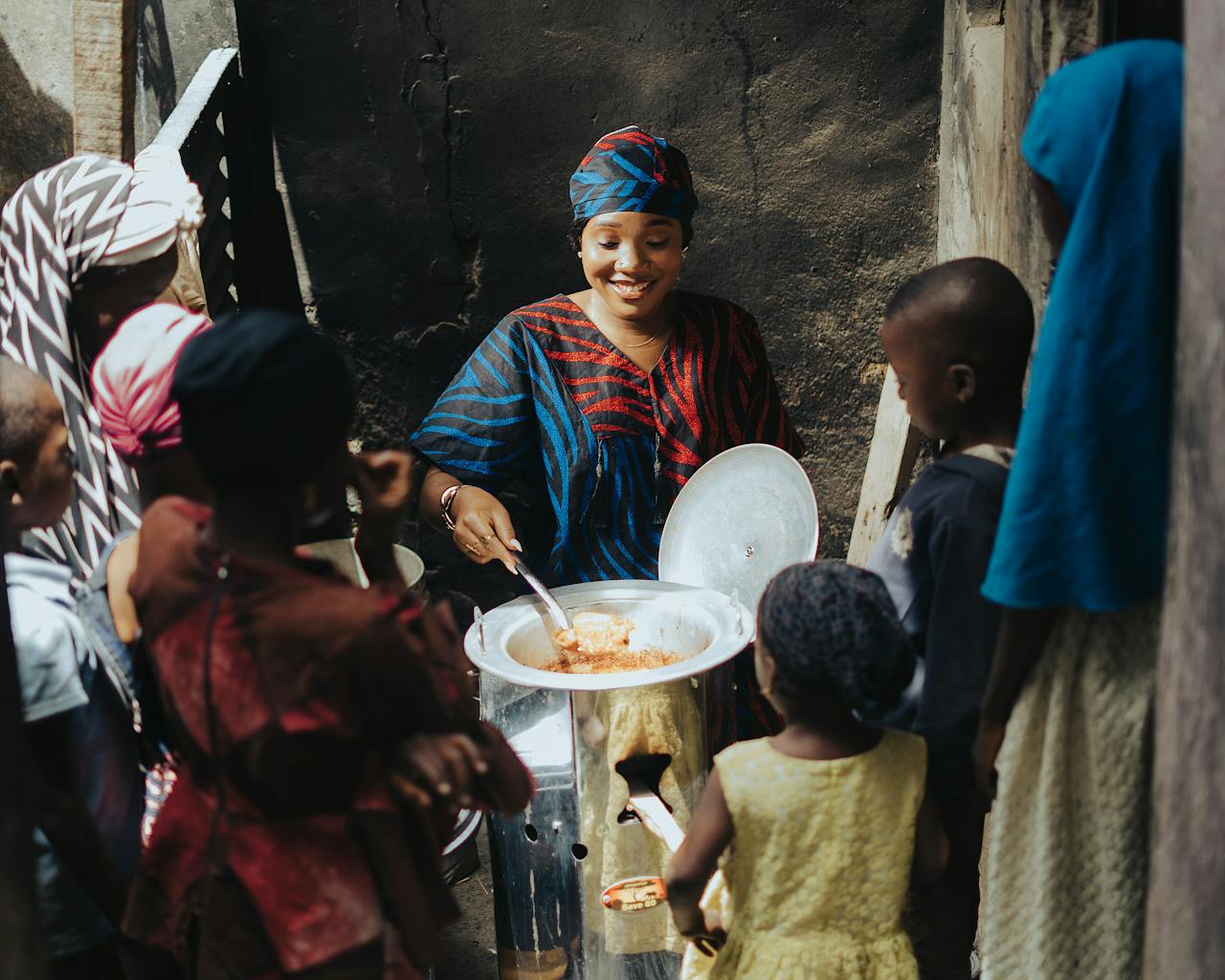 services-03 A woman serves food from a large pot to a group of children outdoors in an African community.
