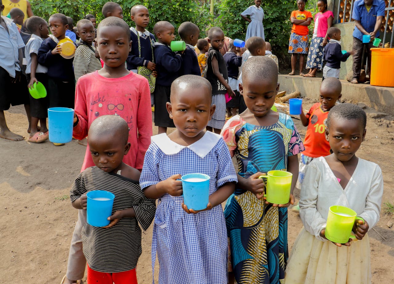 services-02 Group of children holding cups, standing in line outdoors, in a rural setting, waiting for food distribution.