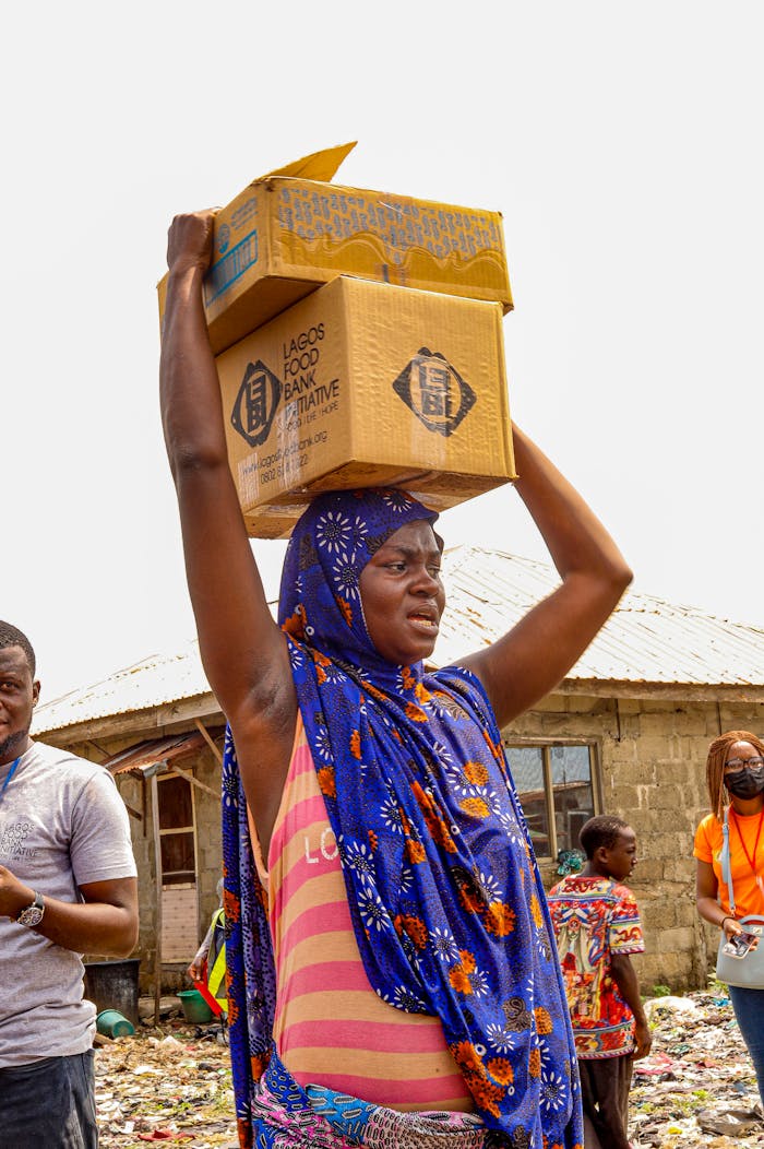 who-we-are African woman in vibrant headscarf carrying boxes in a lively market scene.