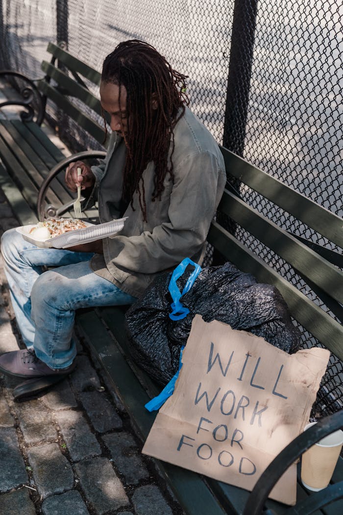 services-01 A homeless man with dreadlocks eating on a park bench beside a 'Will Work For Food' sign.
