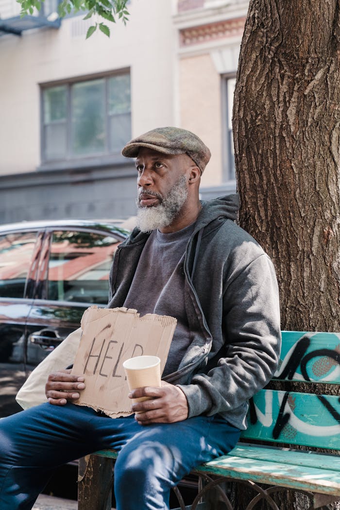 A senior man with a beard sits on a bench holding a sign reading 'Help' in an urban setting.